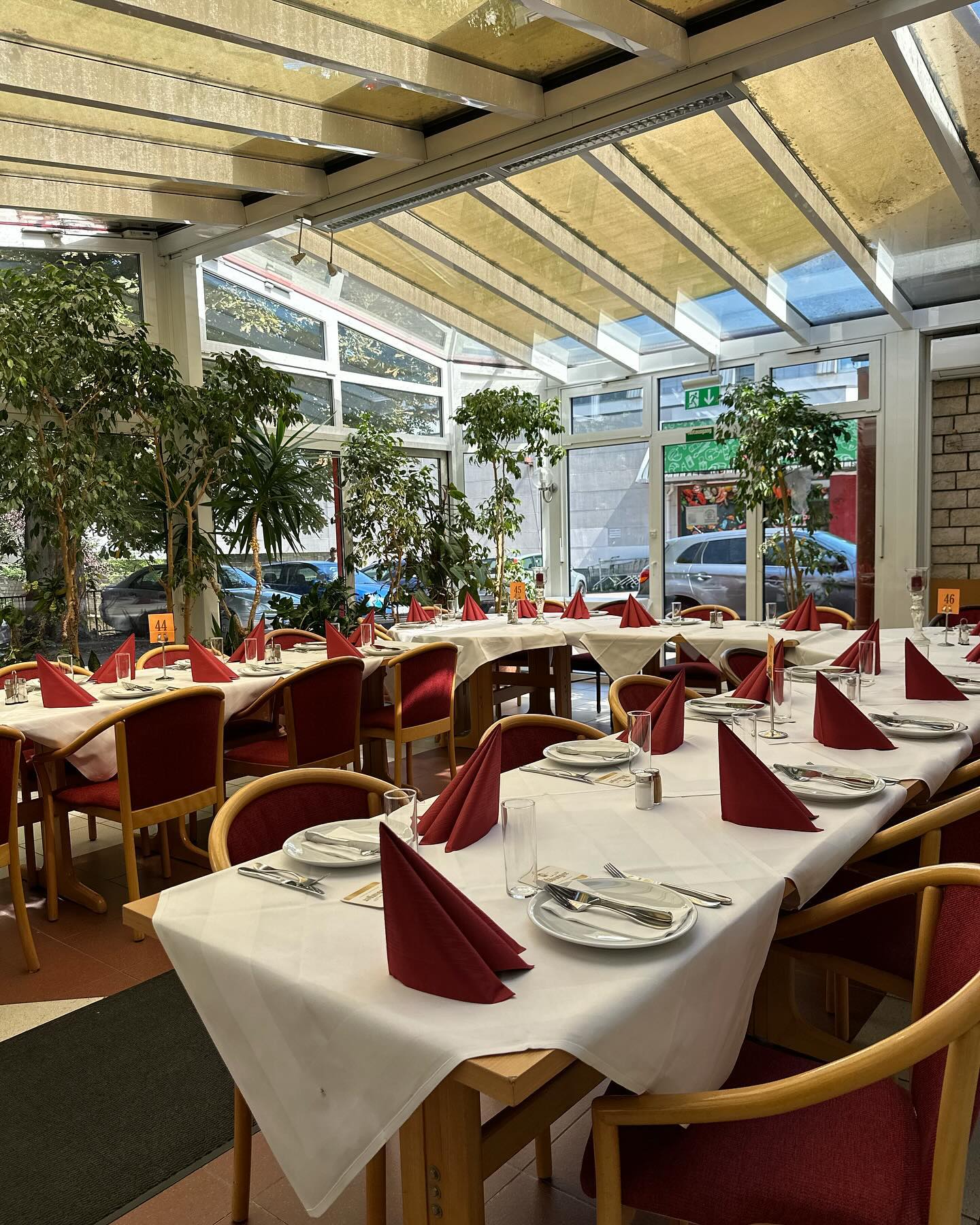 Wintergarten des Steakhaus Haus Wessels: weiß eingedeckte Tische mit bordeauxroten Servietten unter dem Glasdach, umgeben von großen Pflanzen